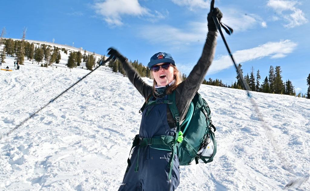 Excited woman in winter gear celebrating on a snowy mountain slope, holding ski poles in the air with a big smile, embodying the joy of backcountry skiing.