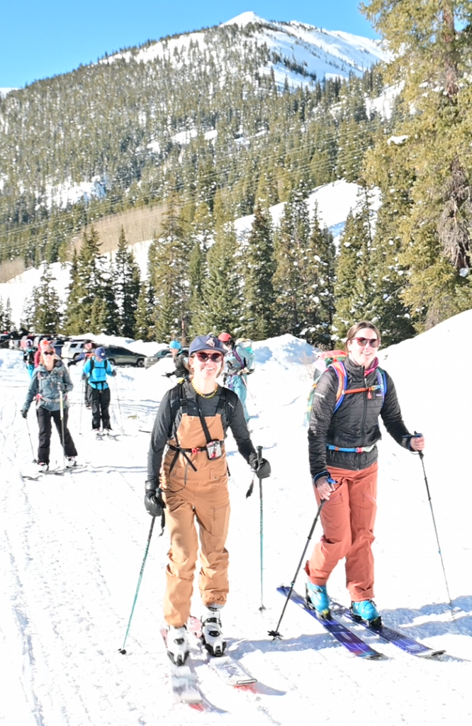 Two smiling women ski touring on a sunny winter day, surrounded by snow-covered mountains and evergreen trees, with a group of backcountry skiers in the background.