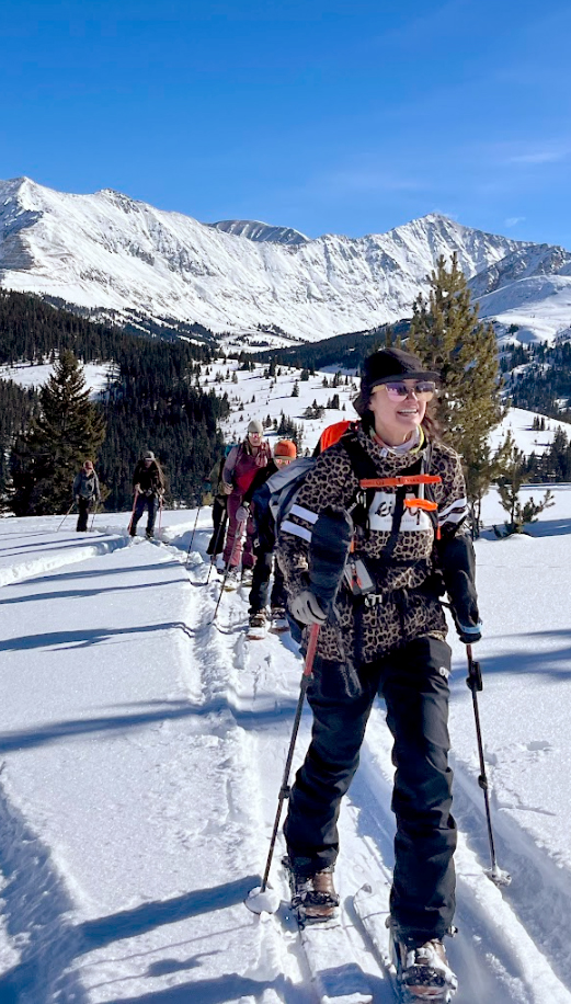 A group of women backcountry skiing under a bright blue sky, led by a smiling skier in leopard-print gear, with snow-covered mountains in the background.