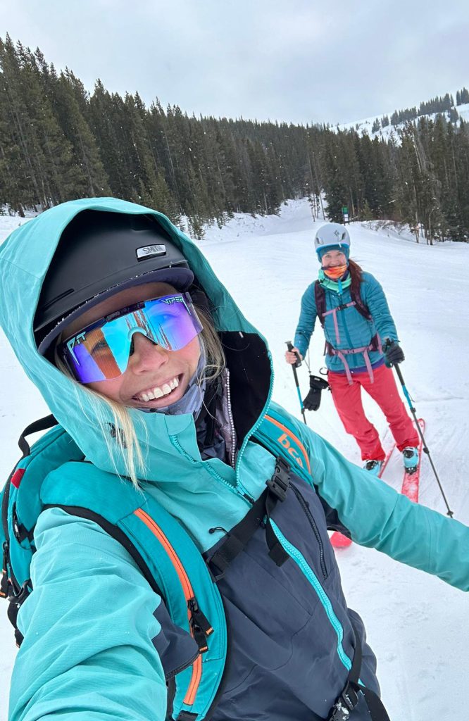 Two women smiling on a backcountry ski tour, dressed in bright turquoise and red winter gear, surrounded by snow-covered trees and mountains.