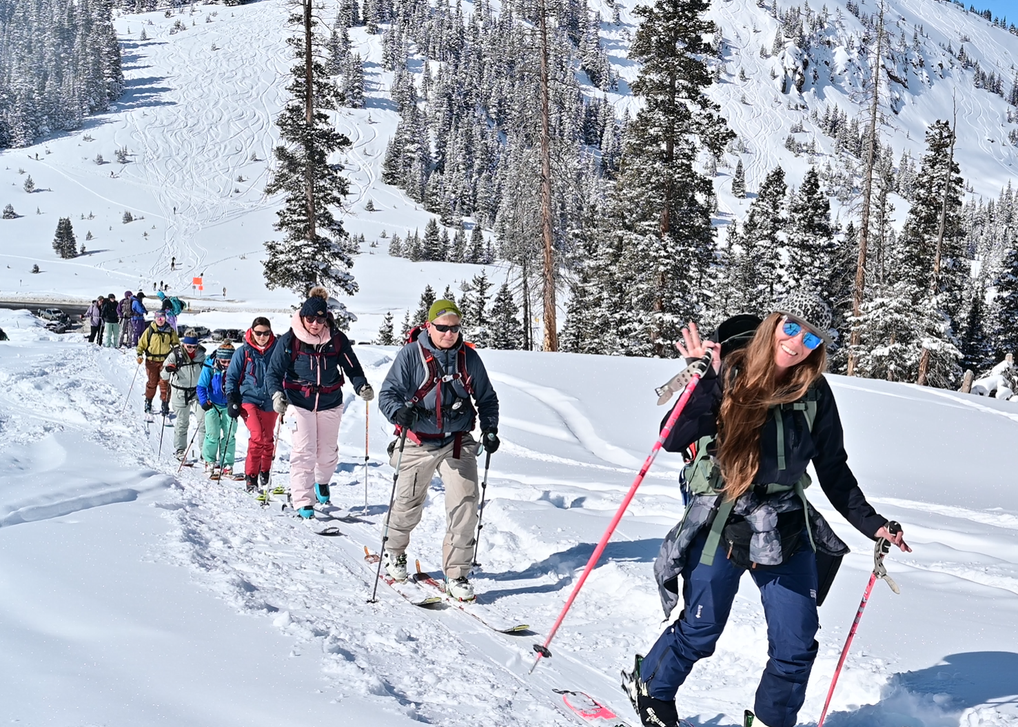 A group of backcountry skiers skinning up a snowy mountain trail, led by a smiling woman waving at the camera, with fresh ski tracks and evergreen trees in the background.
