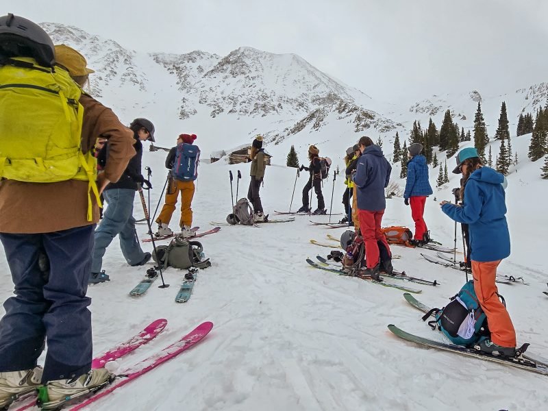 Group of backcountry skiers preparing for an ascent in a snowy alpine setting, surrounded by towering mountains and evergreen trees.