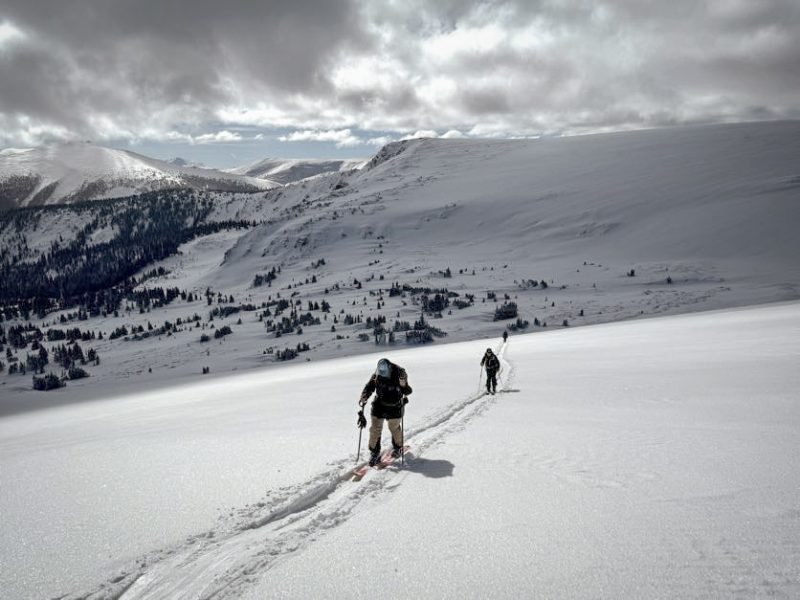 Two backcountry skiers skinning up a vast snow-covered mountain under a dramatic cloudy sky, with untouched powder and alpine scenery stretching into the distance.