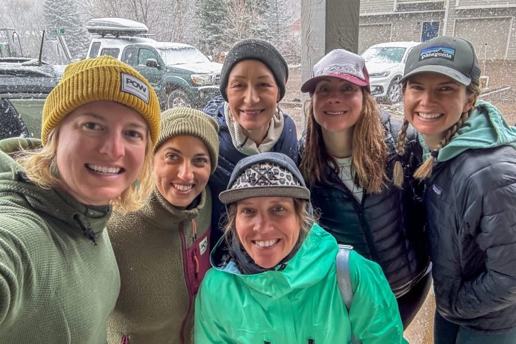 A group of six smiling women dressed in warm outdoor gear, gathered on a snowy day with cars and a cabin in the background.