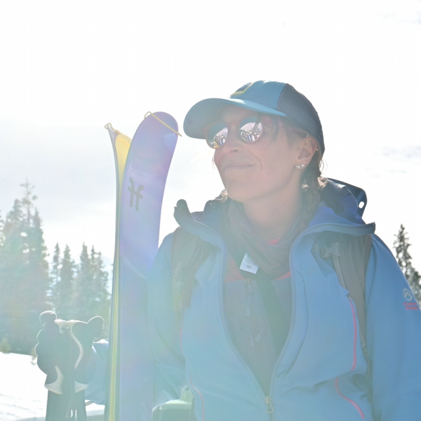 Backcountry skier in a blue jacket and cap, holding skis and poles, smiling in the sunlight with evergreen trees and snow in the background.