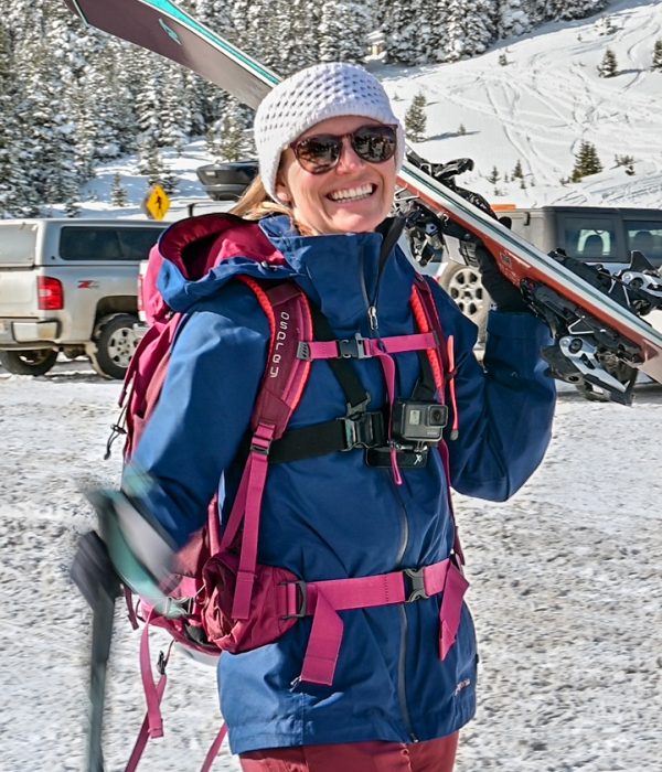 Smiling woman in a blue jacket and pink backpack carrying skis over her shoulder in a snowy parking lot, ready for a backcountry ski tour
