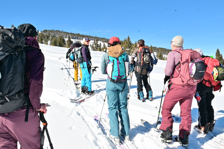 A group of backcountry skiers pausing on a snowy trail under a bright blue sky, dressed in colorful winter gear and carrying backpacks.