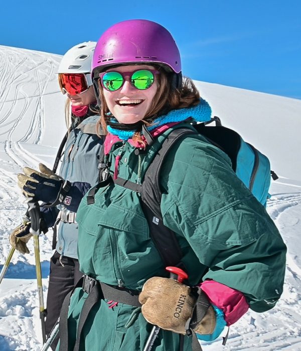 Smiling woman in a green ski suit, pink helmet, and mirrored sunglasses enjoying a bluebird backcountry ski day, with fresh tracks in the snow behind her.