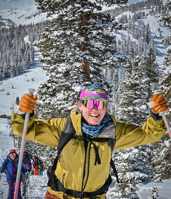 Excited backcountry skier in a yellow jacket and colorful hat skinning uphill with a big smile, surrounded by snow-covered trees and mountains.