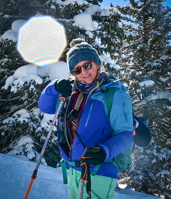 Smiling backcountry skier in a blue jacket and beanie posing with ski poles and an ice axe, surrounded by snow-covered trees on a sunny winter day.